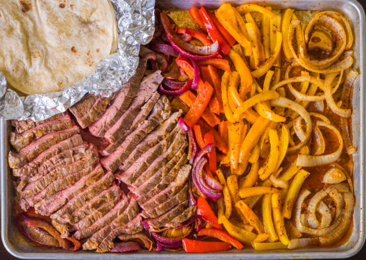 Sheet pan with steak strips, onions, peppers, and tortillas on top