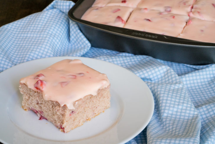 Slice of strawberry sheetcake on a plate with the rest of the sheet cake in the background