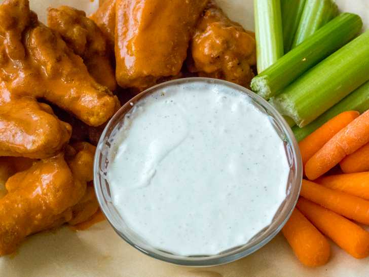 Creamy dip in a bowl surrounded by wings, celary, and carrots