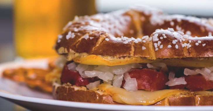 Pretzel bread sandwich with cheese, bratwurst, saurkraut, and sauce.