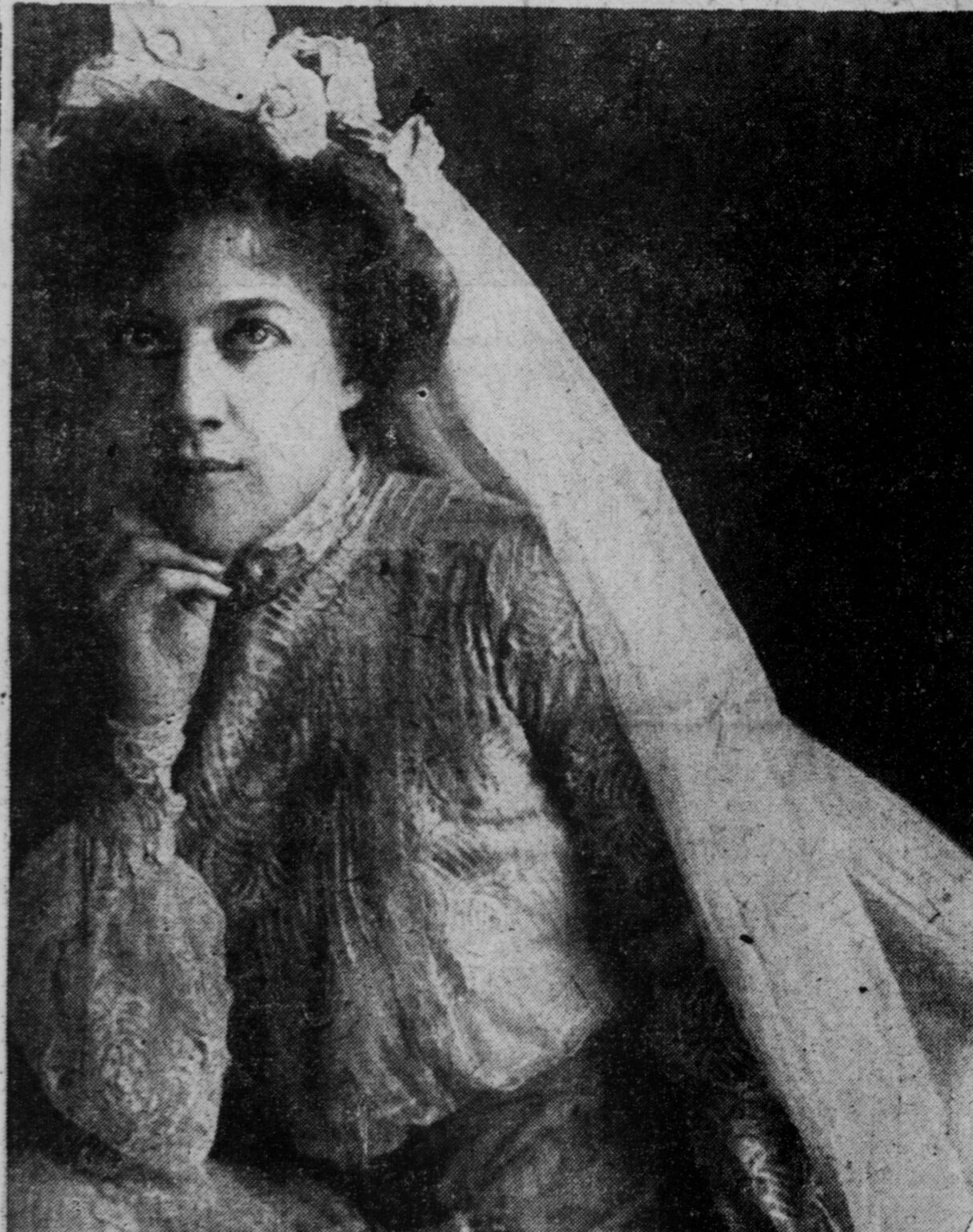 A woman in vintage attire poses thoughtfully, wearing a floral headpiece and veil.