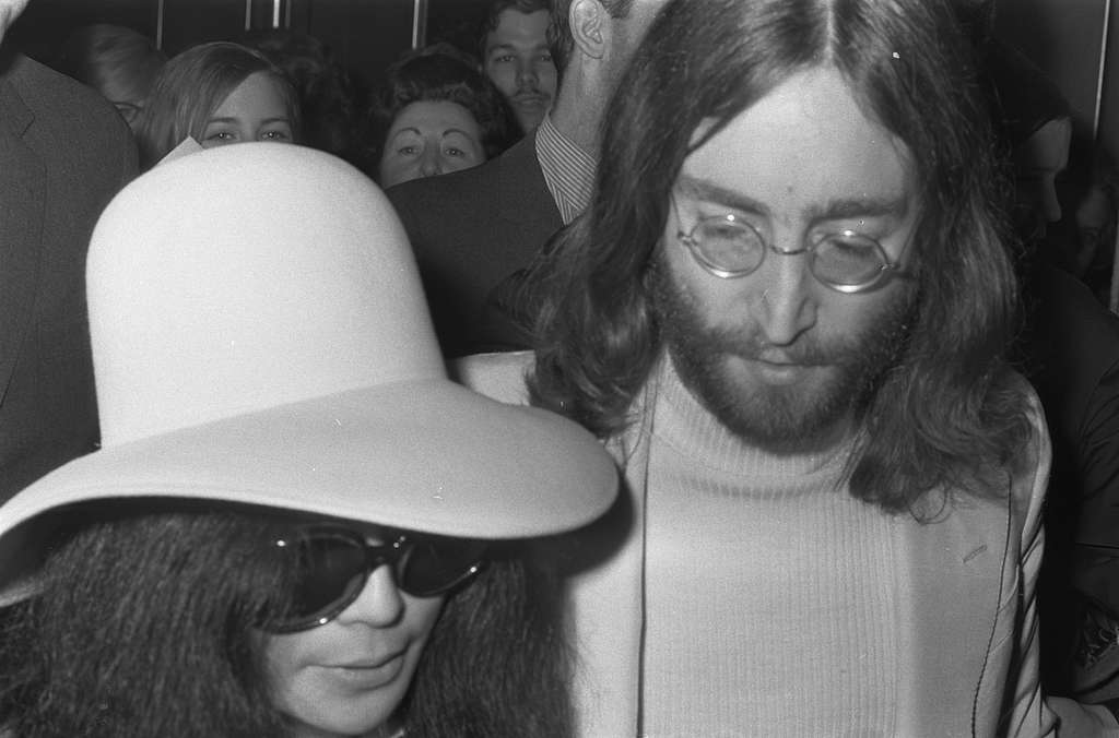 John Lennon on the right with long hair and glasses. Yoko Ono on the left with sunglasses and a sunhat.