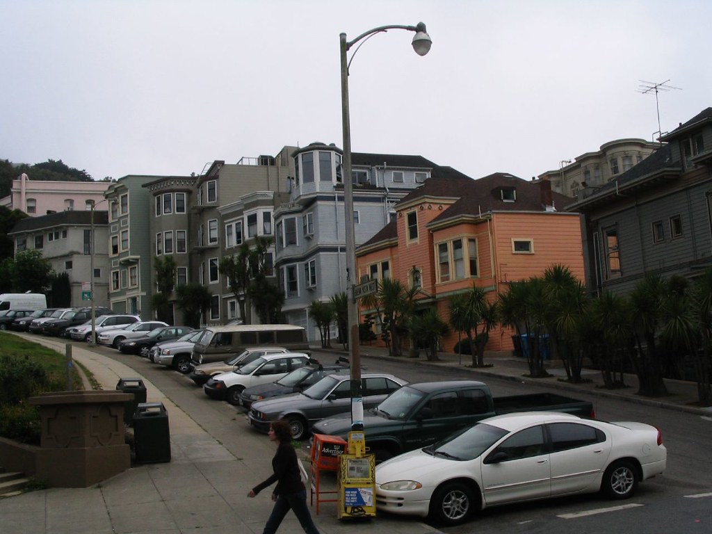 Sloped street with parked cars, a lamppost, and colorful Victorian houses; yellow newspaper box reads “Advertiser.”