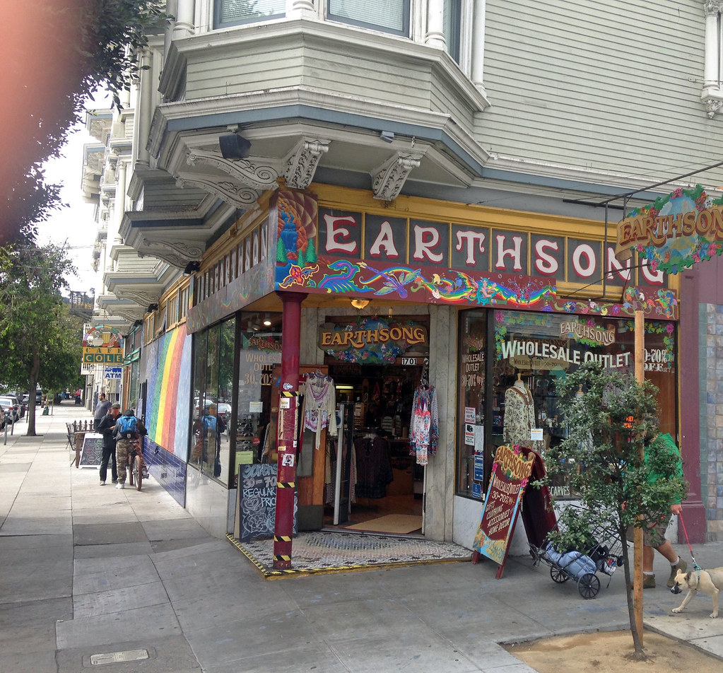 Corner storefront with colorful 'EARTHSONG' sign, 'WHOLESALE OUTLET' in window, rainbow mural; pedestrians and a leashed dog.