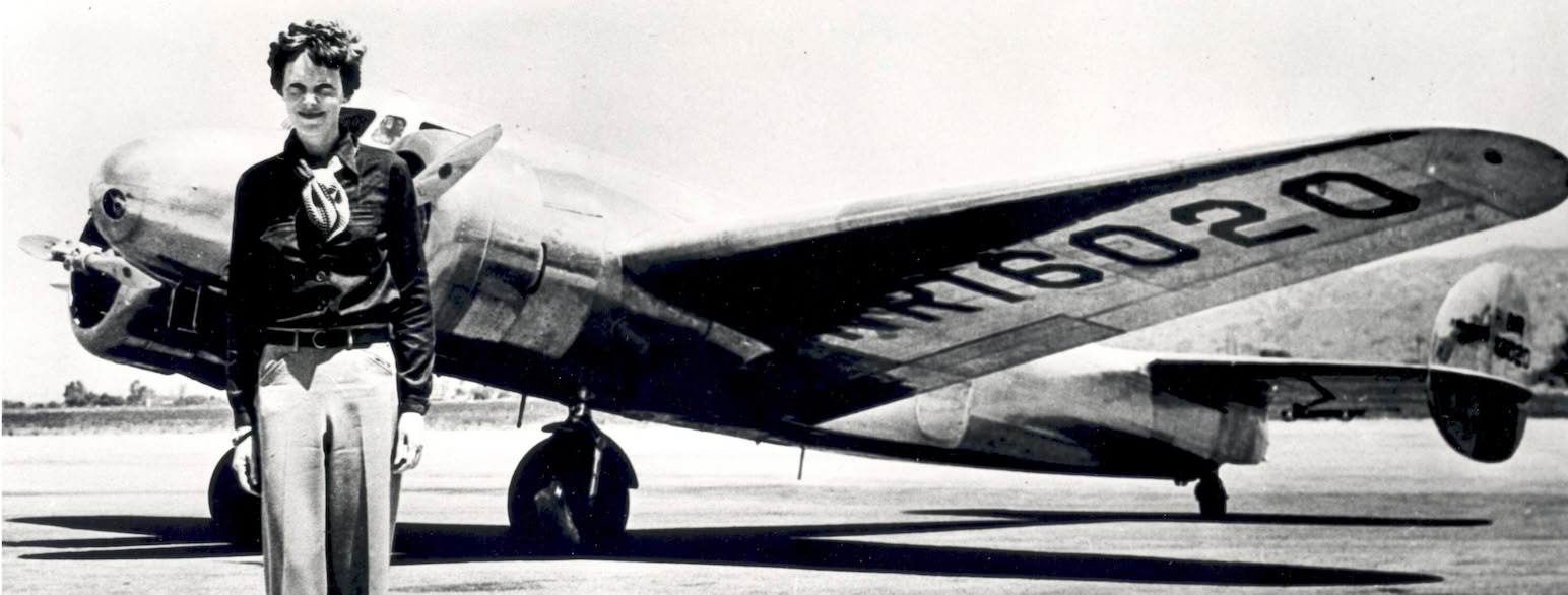 In black and white young Amelia Earhart stands in front of a metal plane. The wing reads: RT6020
