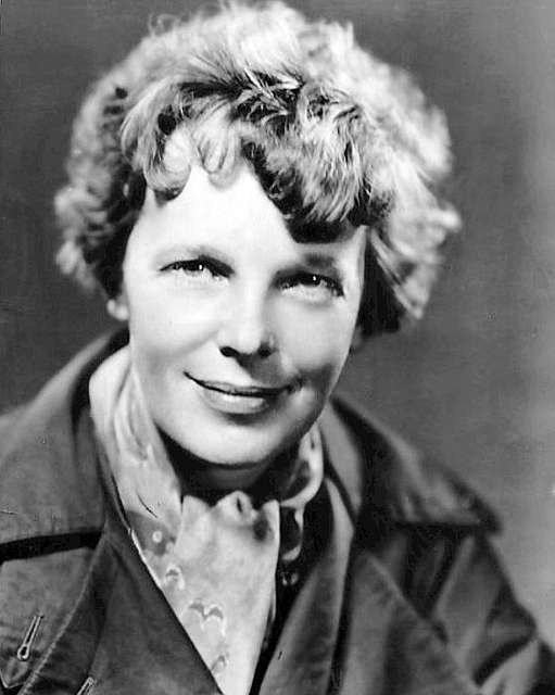 Black and white photo of Amelia Earhart wearing a bandanna around her neck and a jacket. She had short curly hair.