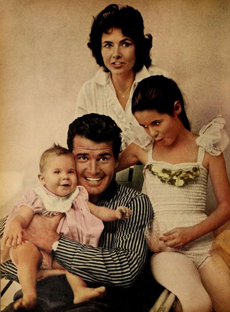 James Garner (center) wears a pinstripe shirt and holds his baby daughter in pink. His other daughter (right) wears a white floral leotard, and his wife stands behind wearing a white shirt.