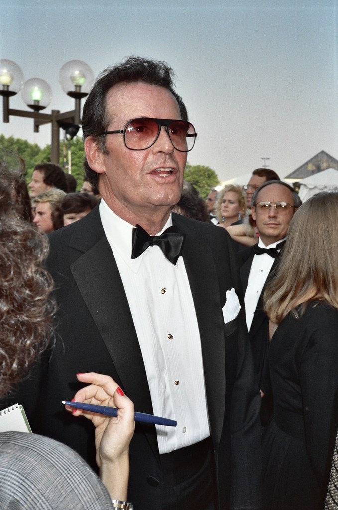An older James Garner wears a black suit with bow tie surrounded by other people at the Emmy awards. He also wears tinted sunglasses and behind him is a large street lamp. The sky is blue.