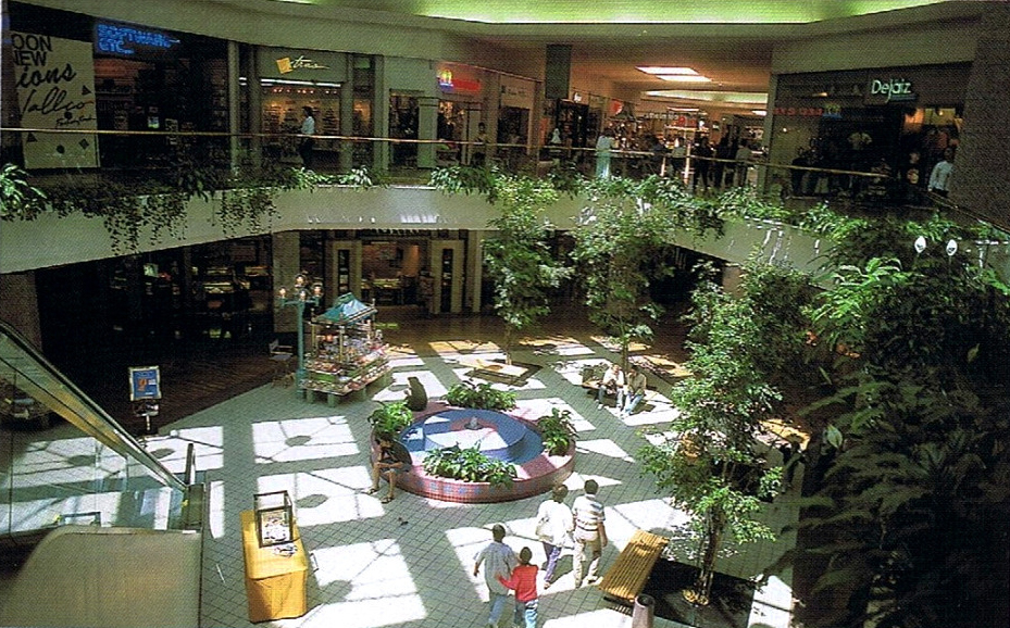 A mall from 1990 with a two stories. On the bottom floor, a circular fountain and many benches provide seats for customers. On the top floor, people walk around from store to store. Many plants hang from the second floor down to the first.