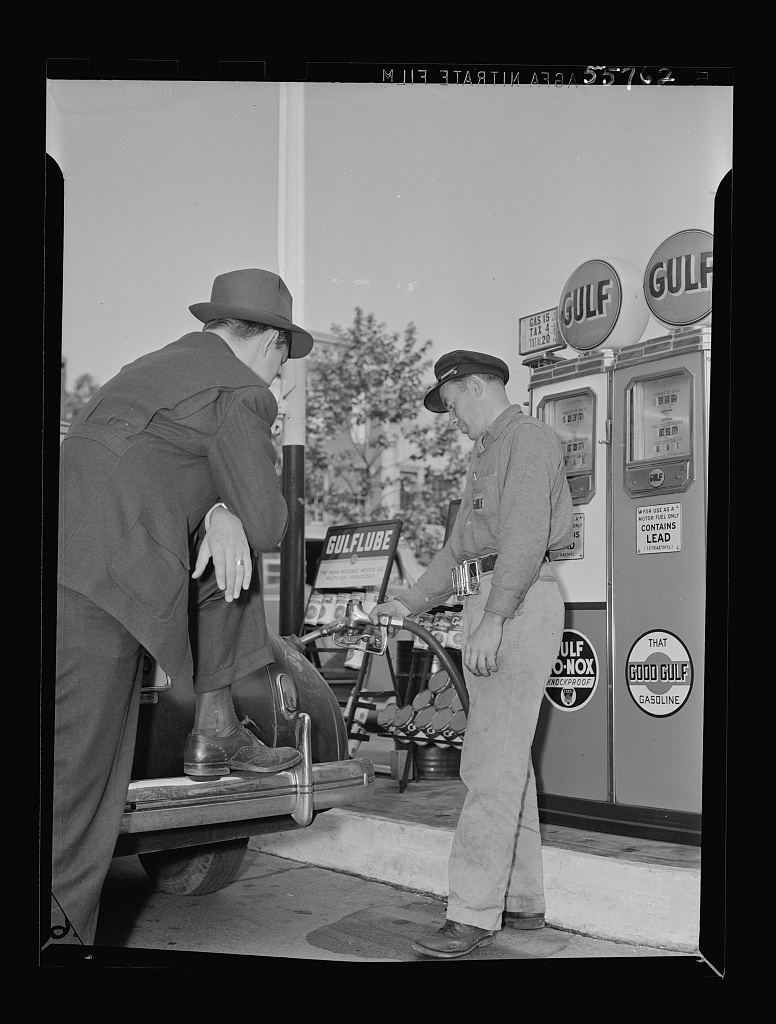 Black and white photo of a man and attendant at a full service gas station. The man wears a black suit and hat and leans on the back of his car. The attendant (right) wears a cap and holds the gas nozzle. Behind him, the pumps read: GULF.