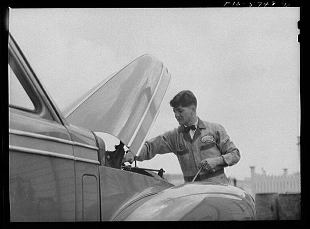 A gas station attendant wearing a bowtie and collared shirt checks the hood of a car.