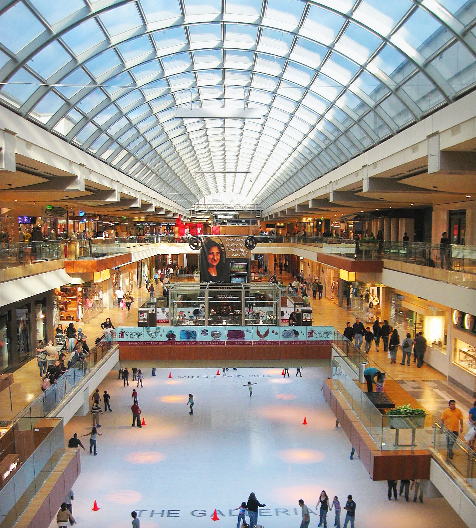 Indoor shopping mall with a glass ceiling featuring an ice skating rink on the lower level and people skating; shops and walkways line both sides above.