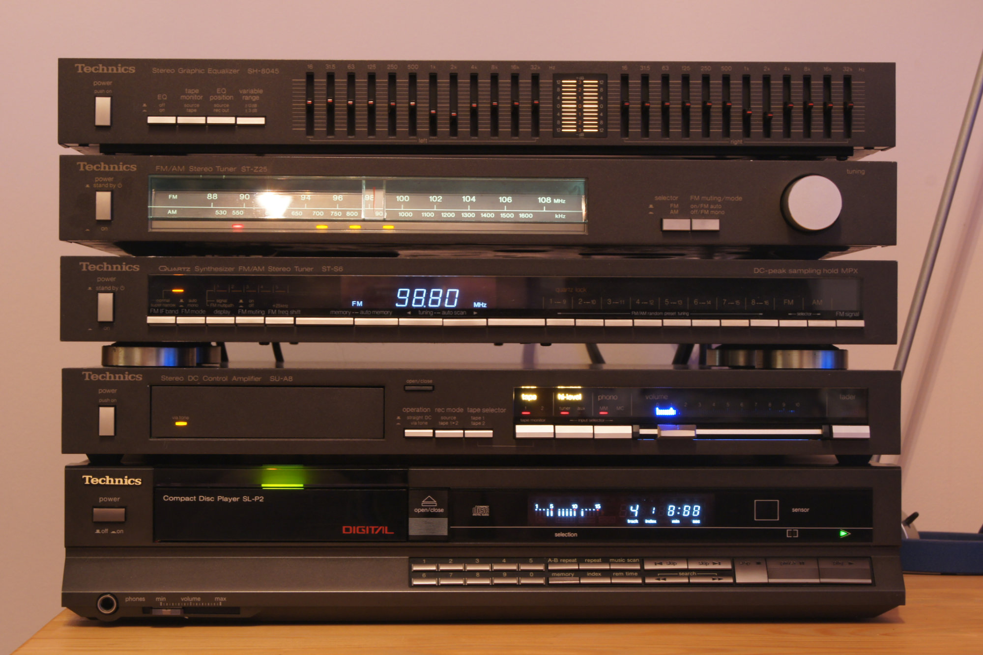 A stack of six vintage Technics audio components, including an equalizer, tuner, amplifier, control amplifier, cassette deck, and CD player, with various display lights illuminated.