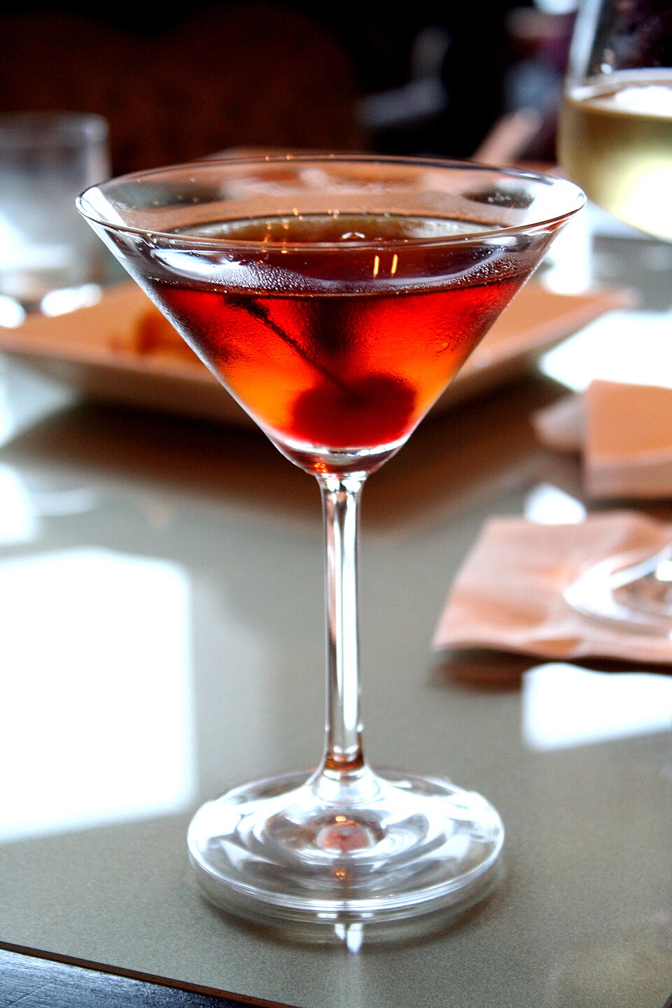 A Manhattan rests in a martini glass. The drink is an amber color and a cherry rests at the bottom of the glass. The glass sits on a grey table top.