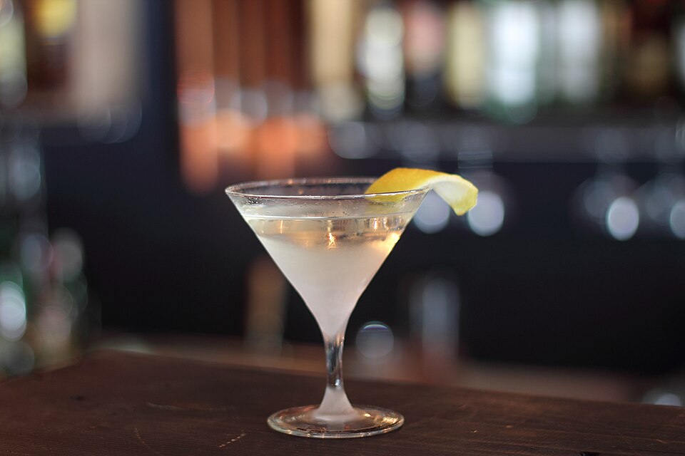 A martini in a small cocktail glass. The glass is frosty cold with a lemon peel as garnish. It rests on a wooden counter top.