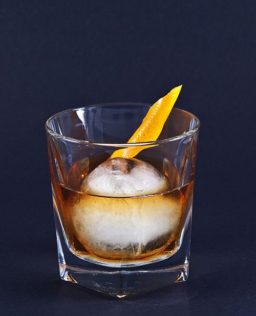 Old Fashioned in a rocks glass. A circular ice cube rests in whiskey. An orange twist on top.