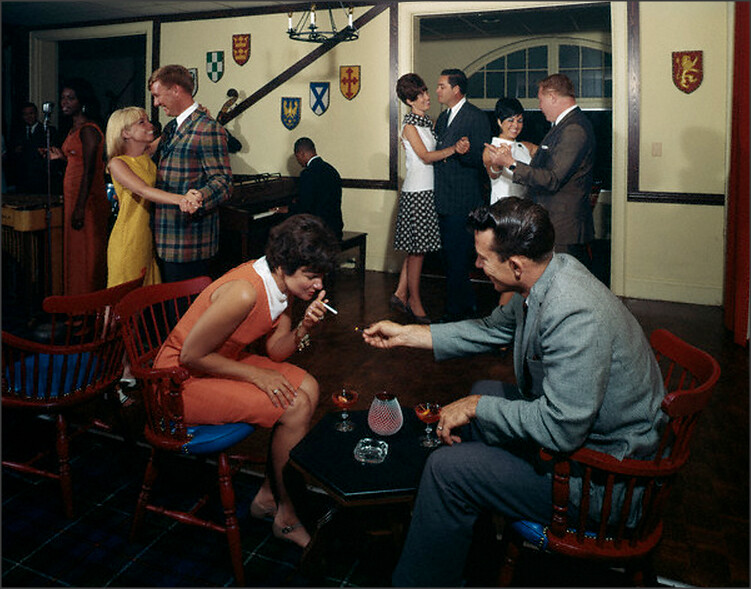 A 1960s party. A couple, woman in orange dress (left) leans forward with a cigarette. Man (right) in a grey suit lights it for her. In between them, a black table holds two manhattans. In the background, couples dance to music performed by a jazz pianist and singer.