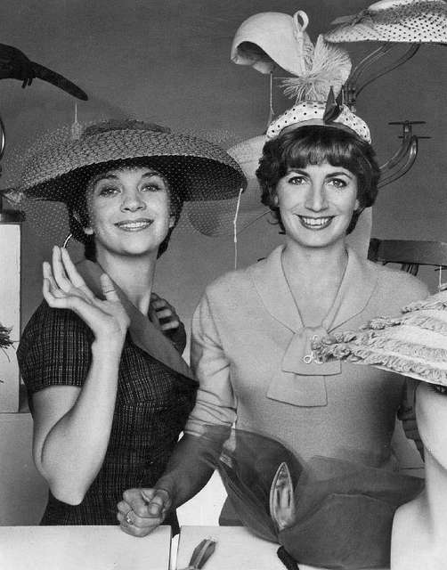 Laverne and Shirley. Left wears a brimmed hat and plaid dress, she waves at the camera. Right wears a dress with a tied neckline and large decorative hat.