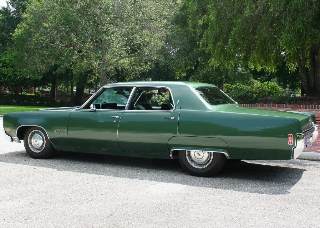 Deep green Oldsmobile Delta 98. In the background is trees and a brick wall.