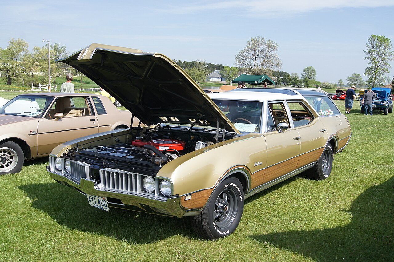 Gold Oldsmobile Vista Cruiser with the hood popped open. It rests on grass and is in front of trees and other cars in a field.