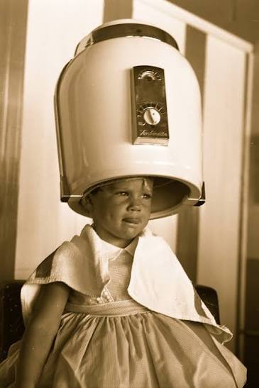 A little girl sits with a bowl around her shoulders in a small chair. Above her head is a huge hooded blow dryer in white.