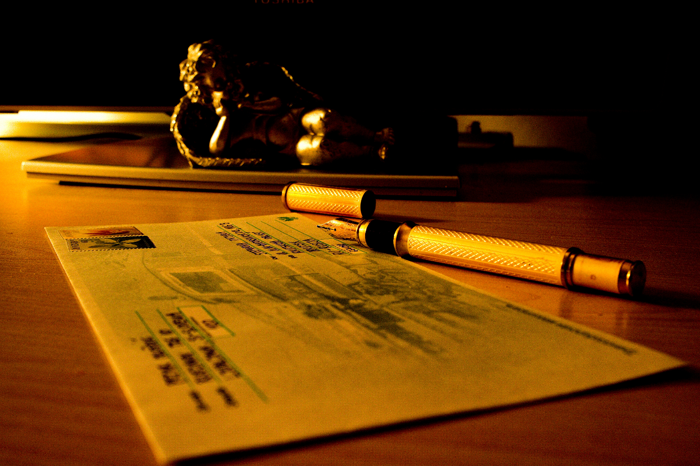 An envelope and calligraphy pen sit on a wooden desk. Behind them is a gold paper weight. The desk glows a yellow/orange color from the desk lamp.