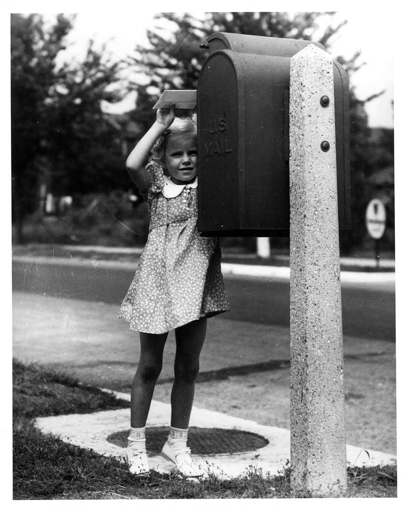 A black and white photo of a girl opening a mailbox and inserting an envelope. The girl is blonde, and wears a floral dress and white shoes.