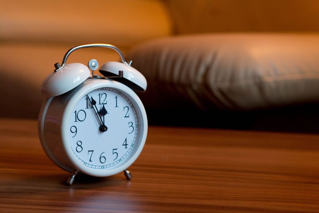 A white alarm clock with black numbers rests on a rustic wooden table, casting a soft shadow.