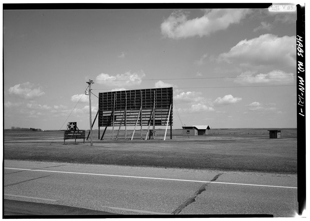 A monochrome photograph capturing a weathered drive-in theater screen, its edges worn, set against a backdrop of trees and soft shadows.