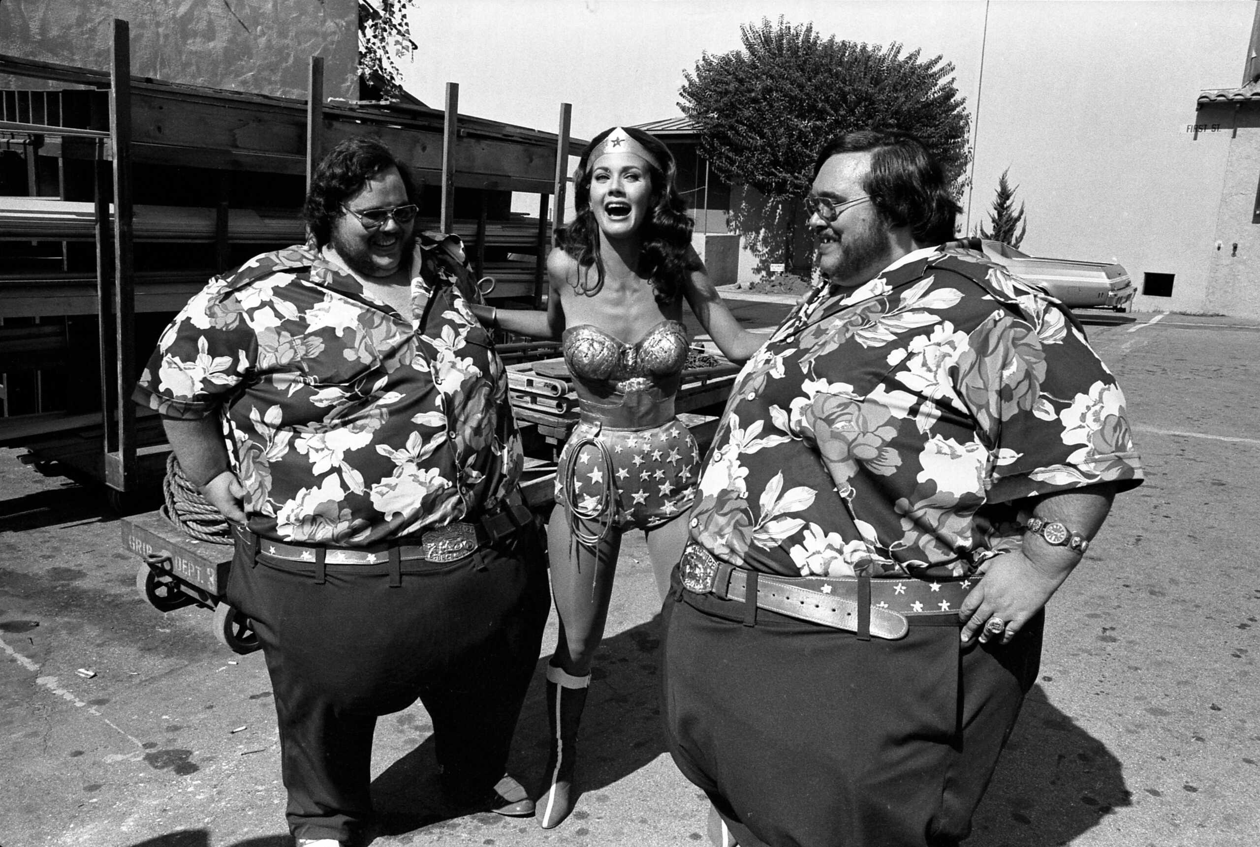 Actors pose playfully together, featuring Wonder Woman amid two larger men in Hawaiian shirts.